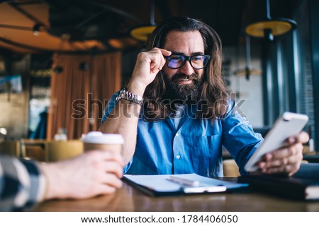 Positive bearded caucasian male using mobile phone for checking banking account owning small business, smiling mature man in spectacles for vision correction satisfied with getting good news via mail