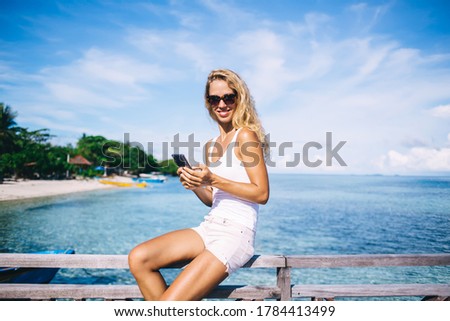 Portrait of cheerful hipster tourist in sunglasses posing at pier with coastline and seashore on background, sincerely female blogger with smartphone technology smiling at camera on Philippines