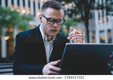 Focused elegant male employee in glasses wearing suit using tablet while sitting on street on blurred buildings and trees background
