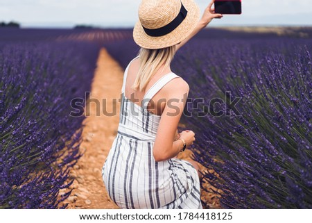 Back view of romantic blond female in hat squatting and making selfie with smartphone during journey through lavender fields in Provence in France