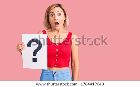 Young blonde woman holding question mark scared and amazed with open mouth for surprise, disbelief face 