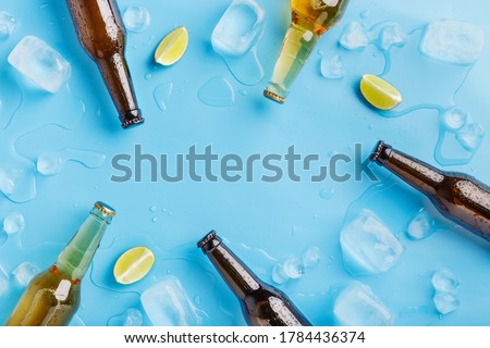 Summer party and chilled drinks. Dark and light glass beer bottles without labels with ice and pieces of lime on blue background, top view, free space