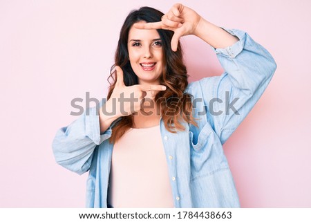 Young beautiful brunette woman wearing casual clothes over pink background smiling making frame with hands and fingers with happy face. creativity and photography concept. 