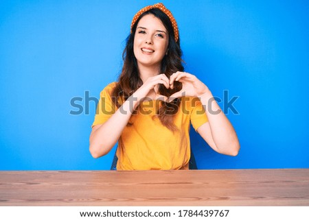 Young beautiful brunette woman sitting on the table over blue background smiling in love showing heart symbol and shape with hands. romantic concept. 