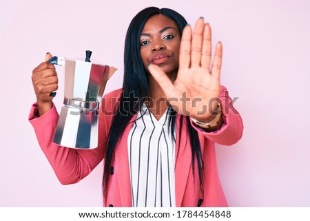 Young african american woman holding italian coffee maker with open hand doing stop sign with serious and confident expression, defense gesture 