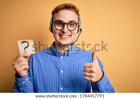 Redhead call center agent man working using headset holding reminder with question mark Smiling happy and positive, thumb up doing excellent and approval sign