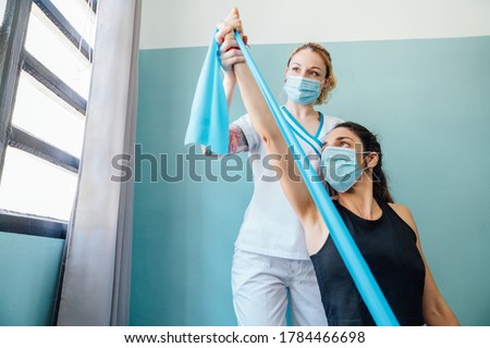 Professional Caucasian physical therapy woman at a clinic giving a Pilates stretch treatment with a rubber band to a client with a face mask due to the covid 19 coronavirus pandemic. Royalty-Free Stock Photo #1784466698