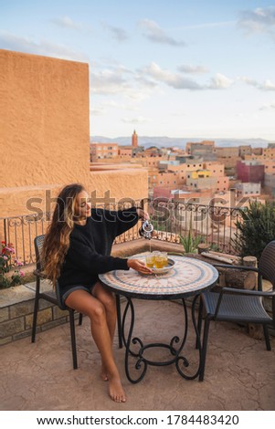 Woman pouring traditional moroccan mint tea on terrace with amazing view of old arab town Boumalne in Morocco at sunset. Round vintage table.