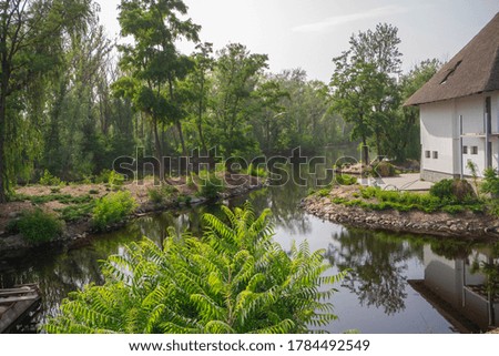 A horizontal photo of a river flowing past a house with white walls and a shore with tall green trees. Daytime. Clear sky. Landscape