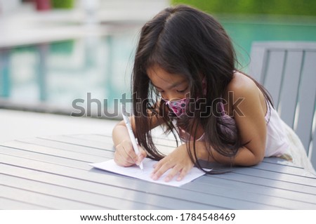 Portrait of a cute 4-5 year old Asian girl Drawing in the outdoor garden and wearing a mask.