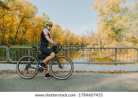 Young male cyclist in helmet and sportswear stands in park on background of autumn landscape with river and looks into camera. Athlete cyclist walking in the forest on a bicycle.