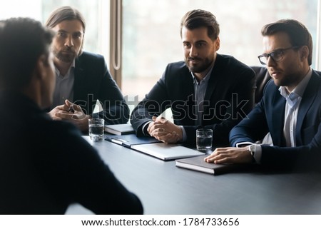 Focused young male group of employers in formal suits sitting at table with vacancy candidate, involved in job interview in office. Attentive confident hr managers listening to seeker at meeting. Royalty-Free Stock Photo #1784733656