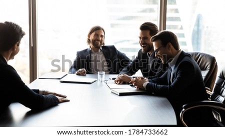 Team of male employers laughing at job interview with male candidate, enjoying relaxed humorous atmosphere in office. Happy businessmen having fun at negotiations meeting, business relations concept.
