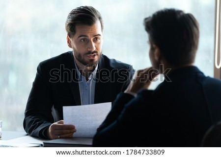 Head shot serious young businessman in formal suit holding paper cv resume in hands, discussing contract details with job candidate or checking working experience at interview meeting in office.