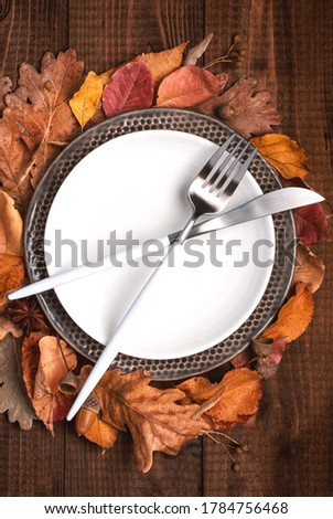 White plate, cutlery and autumn leaves on a dark brown wooden background top view, flat lay. 