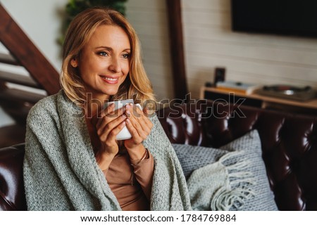Close up of dreaming middle aged woman sitting in living room with cup of coffee or tea enjoying under blanket Royalty-Free Stock Photo #1784769884