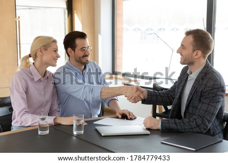 Excited young Caucasian couple handshake greeting with male real estate agent at meeting in office, happy husband shake hand close deal make agreement with broker, ownership, acquaintance concept