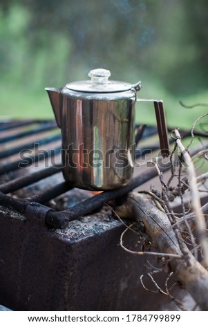 Stainless steel coffee carafe percolator on campfire grate with stick.