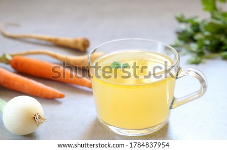 Clear homemade bone  broth in the glass cup on the table. Carrot,onion and parsley are around. Horizontal orientation.