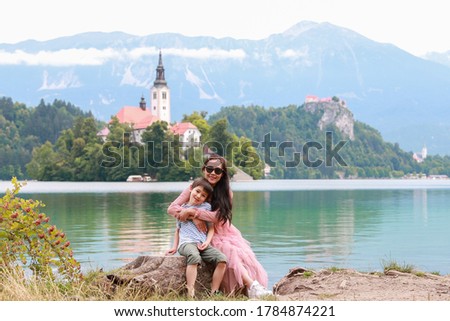 Young tourist mother hugging her son next to lake bled with beautiful Pilgrimage Church of the Assumption of Maria and bled castle on background. Asian woman travel in Slovenia with family.