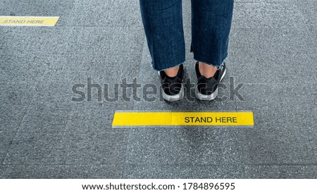 Woman feet standing on floor with STAND HERE text on yellow line print for social distancing. Public Coronavirus safety