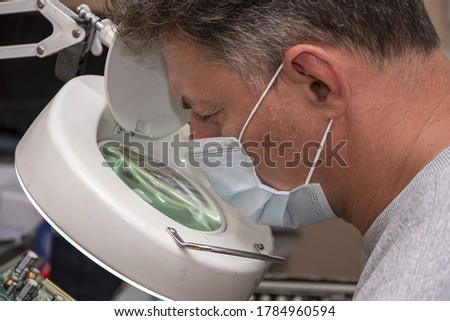 A 40-45-year-old man in a medical mask looks through a magnifying glass at an electrical Board from a computer. Repair of office equipment at home.