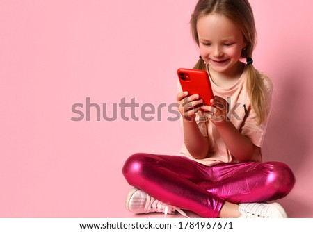 Blonde little girl in t-shirt, leggings and sneakers. She smiling, using her red smartphone while sitting on floor with crossed legs against pink background. Modern technologies.