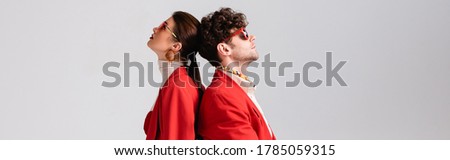 panoramic shot of trendy couple in red blazers and sunglasses standing back to back isolated on grey Royalty-Free Stock Photo #1785059315