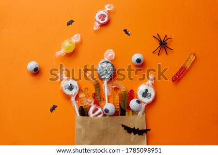 halloween, sweets and holiday concept - candies in paper bag and decorations on orange background