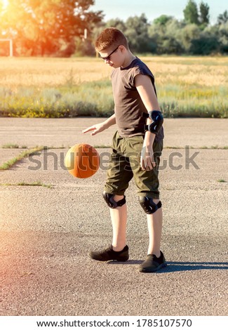 Caucasian teenage boy in a black T-shirt and green shorts plays with a basketball on a summer playground. Basketball player training. Health and Sports. Sunset, bokeh. People healthy lifestyle concep