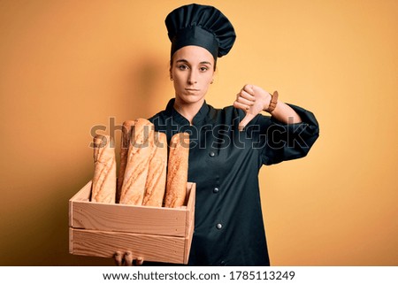 Young beautiful baker woman wearing cooker uniform and hat holding wooden box with bread with angry face, negative sign showing dislike with thumbs down, rejection concept