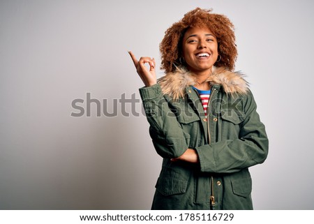 Young beautiful African American woman with curly hair wearing green casual winter coat with a big smile on face, pointing with hand and finger to the side looking at the camera.