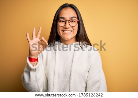 Young beautiful asian woman wearing casual sweater and glasses over yellow background smiling positive doing ok sign with hand and fingers. Successful expression.