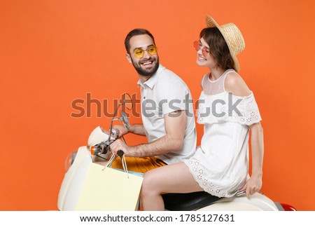 Side view of smiling young couple friends guy girl in hat glasses driving moped isolated on orange wall background. Driving motorbike transportation concept. Hold package bag purchases after shopping