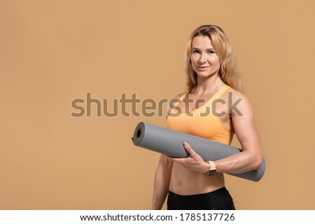 Pilates, fitness and yoga in middle age. Friendly woman in sportswear with fitness tracker and mat, isolated on yellow background, studio shot
