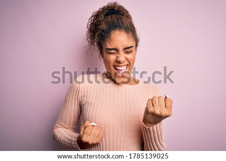 Young beautiful african american girl wearing casual sweater standing over pink background celebrating surprised and amazed for success with arms raised and eyes closed. Winner concept.