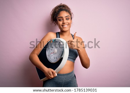 Young african american sporty girl holding weighing machine over isolated pink background happy with big smile doing ok sign, thumb up with fingers, excellent sign