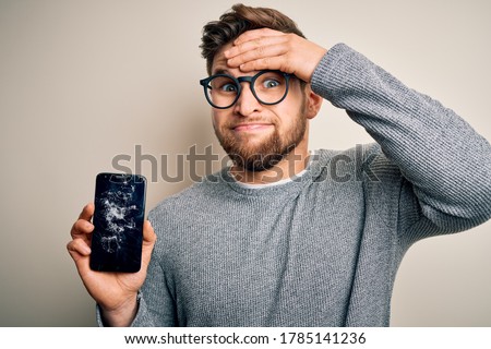 Young blond man with beard and blue eyes wearing glasses holding broken smartphone stressed with hand on head, shocked with shame and surprise face, angry and frustrated. Fear and upset for mistake.