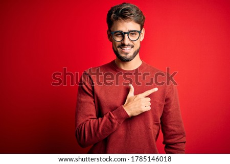 Young handsome man with beard wearing glasses and sweater standing over red background cheerful with a smile on face pointing with hand and finger up to the side with happy and natural expression