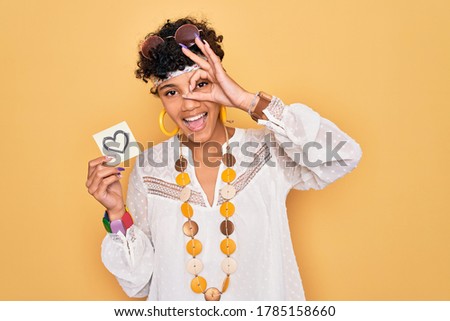 Beautiful african american afro hippie woman wearing sunglasses holding reminder with heart with happy face smiling doing ok sign with hand on eye looking through fingers