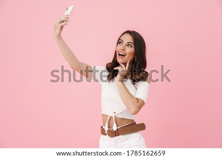 Image of joyful attractive woman taking selfie on cellphone and smiling isolated over pink background