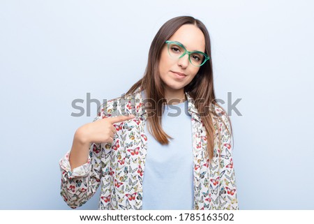 young pretty woman looking proud, confident and happy, smiling and pointing to self or making number one sign against blue wall