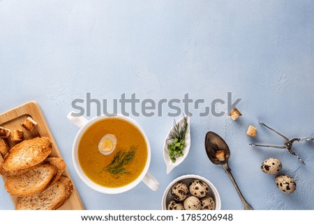 Chicken bouillon with dill and quail egg in bowl, baguette toasts and croutons on cutting board, spoon and tongs. Flat lay on blue background. Large copy space. Healthy eating and dieting concept.