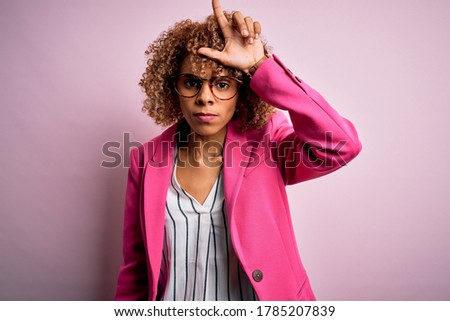 Young african american businesswoman wearing glasses standing over pink background making fun of people with fingers on forehead doing loser gesture mocking and insulting.