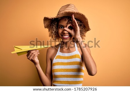 African american tourist woman with curly on vacation wearing summer hat holding paper plane with happy face smiling doing ok sign with hand on eye looking through fingers