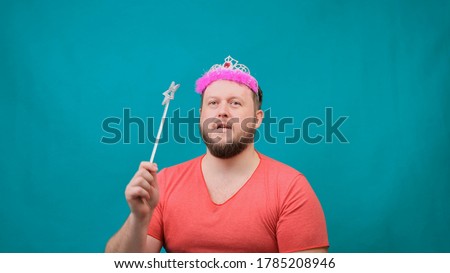 funny bearded freaky man in a pink T-shirt with a deadema on his head is dreaming with a magic wand in his hand. A funny wizard joke to make and fulfill a wish