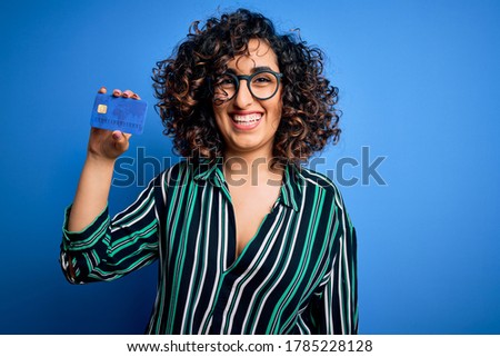 Young beautiful curly arab woman wearing glasses holding credit card money for payment with a happy face standing and smiling with a confident smile showing teeth