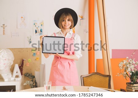 Young smiling pretty girl artist in hat and apron showing blank screen tablet in workshop. Cozy photo for ad or blog