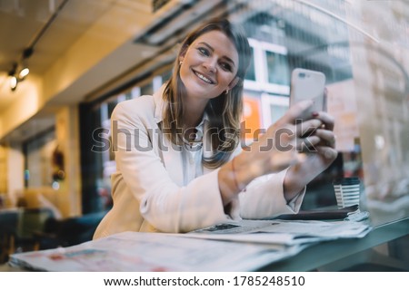 Happy Caucasian woman smiling at front camera while clicking selfie content pictures, cheerful hipster girl enjoying vlog streams using cellular device for shooting web video during pastime in cafe