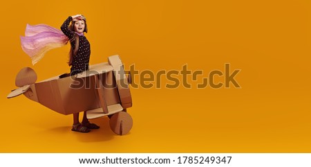 Cute dreamer girl playing with a cardboard airplane. Childhood. Fantasy, imagination. Studio portrait on a yellow background. Royalty-Free Stock Photo #1785249347